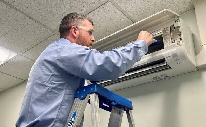 HVAC technician on stepladder performing delicate maintenance and coil cleaning on wall-mounted air handler